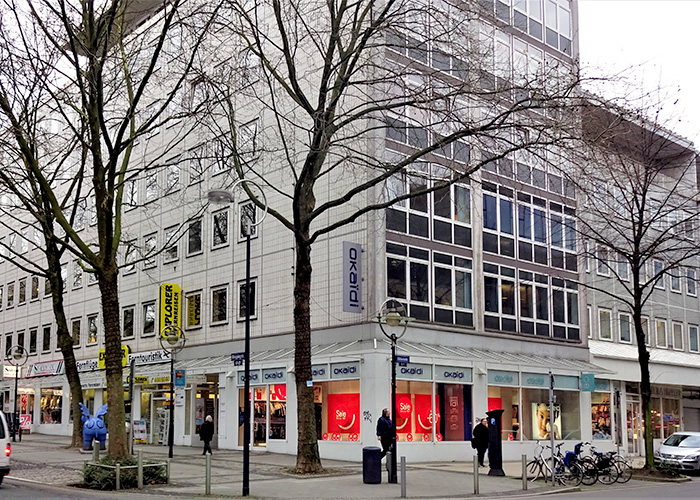 Großes Gebäude mit Ladenlokalen im EG, Bäume und Fussgänger