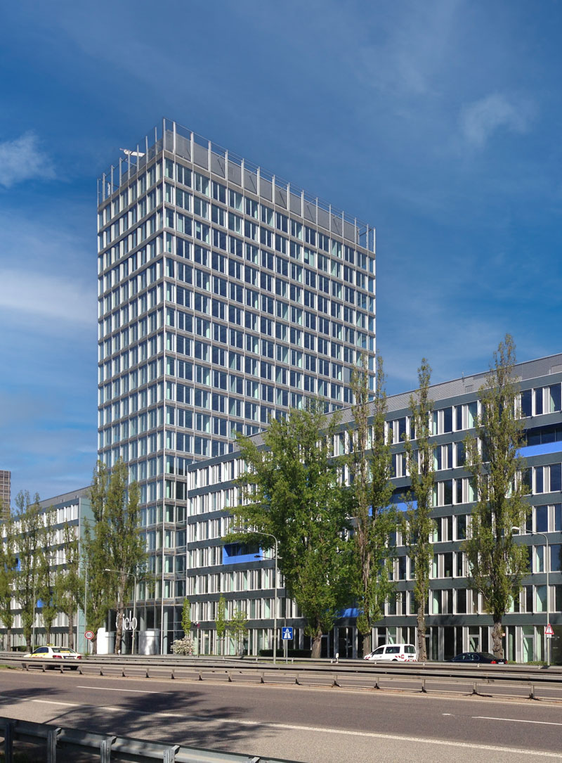 großes, mehrstöckiges Gebäude, Straße und hohe Bäume im Vordergrund
