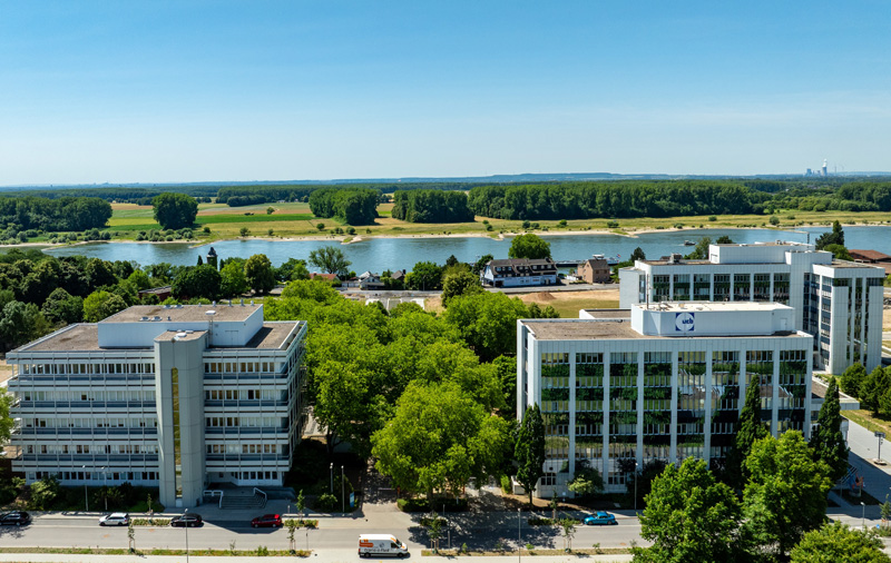 Blick von oben auf drei Bürogebäude mit Glasfassade und viele Bäumen, im Hintergrund der Rhein